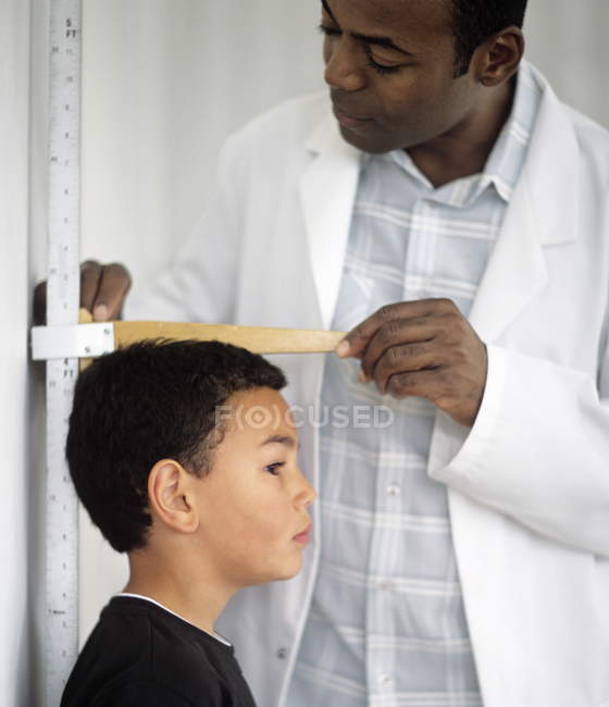 African American doctor checking height of elementary age boy with measuring equipment. — Stock Photo