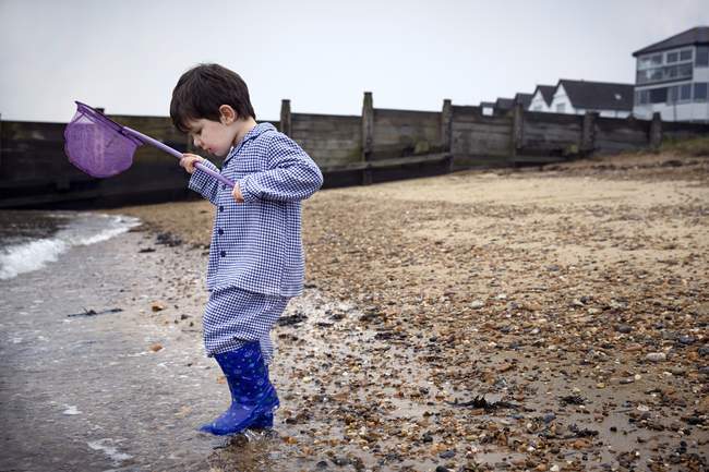 Preschooler boy in rubber boots on beach holding fishing net. — Stock Photo