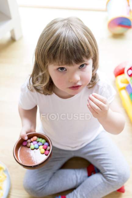 Vorschulmädchen sitzt mit Schale voller Süßigkeiten auf dem Boden. — Stockfoto