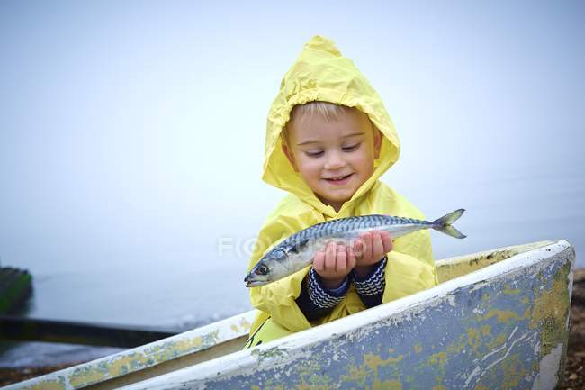 Toddler boy in yellow raincoat holding mackerel fish in boat. — Stock Photo