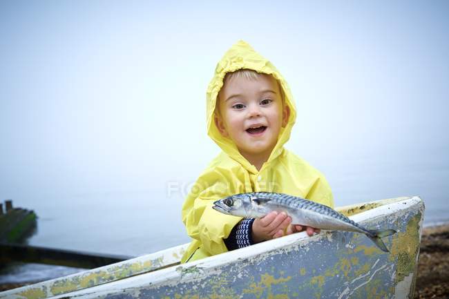 Хлопчик Тоддлер у жовтому плащі тримає макрельську рибу в човні . — стокове фото