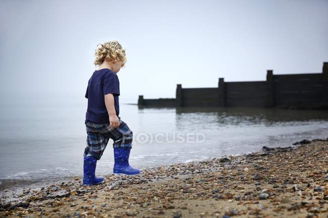 Хлопчик в гумових чоботях, що ходять на піщаному пляжі . — стокове фото
