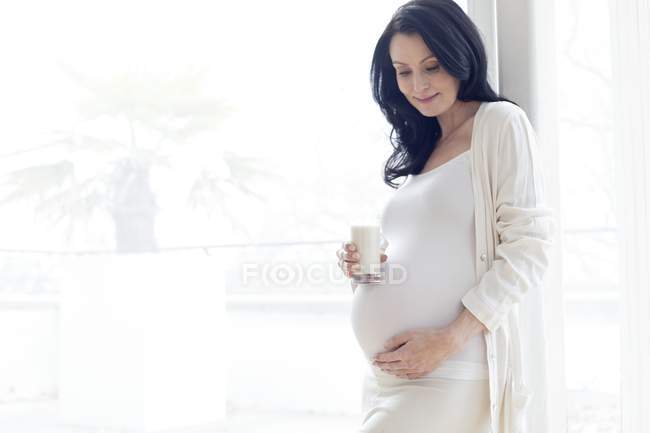Mujer embarazada tocando la barriga mientras sostiene un vaso de leche . - foto de stock