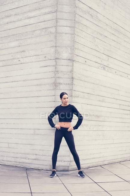Donna in abbigliamento sportivo in piedi in un angolo di edificio con le mani sui fianchi . — Foto stock