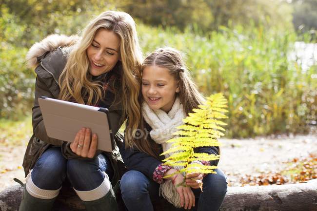 Mother using digital tablet with daughter on log in forest. — Stock Photo