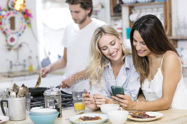 Las mujeres usando smartphone en la cocina mientras desayunan . - foto de stock