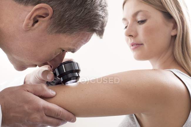 Médico masculino examinando toupeira no braço de mulher jovem, usando um dermatoscópio . — Fotografia de Stock
