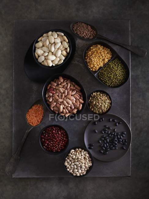 Legumes in bowls on dark background, overhead view. — Stock Photo