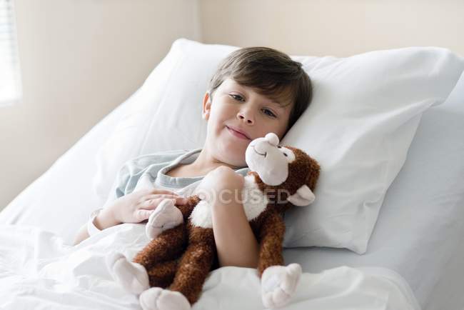 Boy lying with stuffed monkey in hospital bed. — Stock Photo
