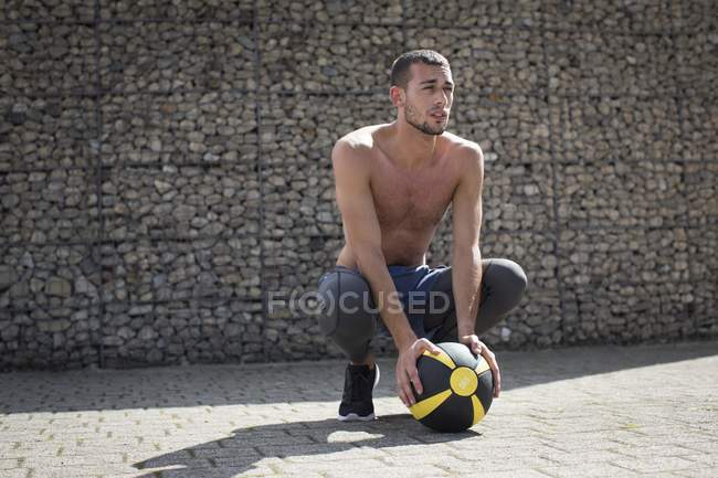 Mann kauert mit Medizinball vor Steinmauer. — Stockfoto