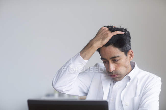 Stressed doctor with hand on forehead looking down at laptop. — Stock Photo