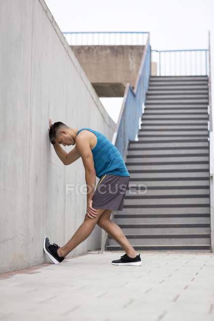 Young man stretching leg against wall of street stairs. — Stock Photo