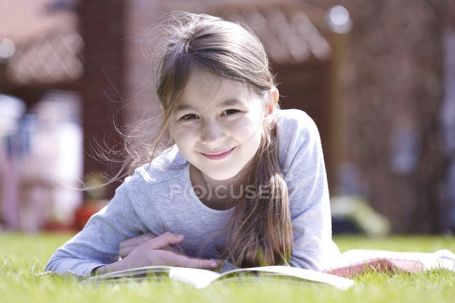 Preadolescente chica acostada en la manta en el prado verde con libro y mirando en la cámara . - foto de stock