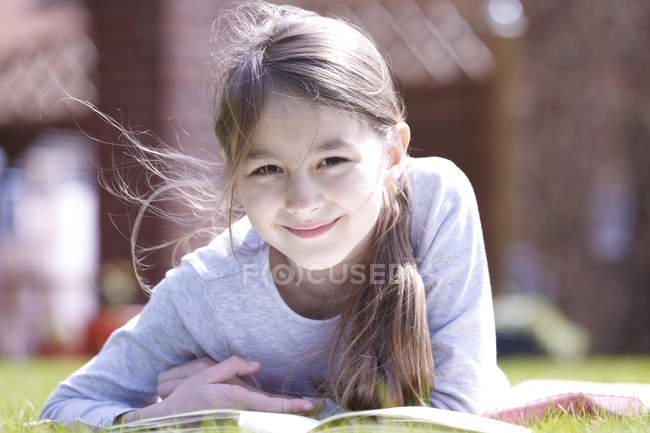 Preadolescente chica acostada en la manta en el prado verde con libro y mirando en la cámara
. - foto de stock
