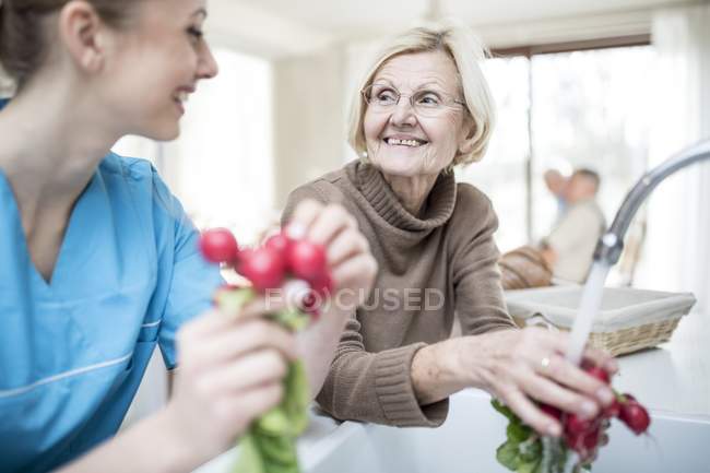Female care worker washing radishes with senior woman in care home. — Stock Photo