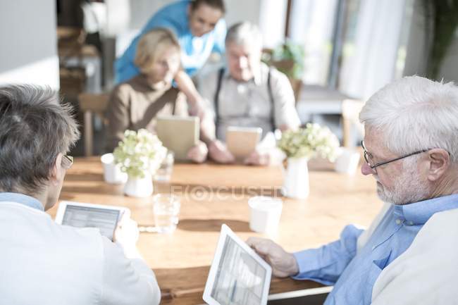 Adultos idosos usando computadores tablet à mesa com enfermeira ajudando na casa de cuidados . — Fotografia de Stock