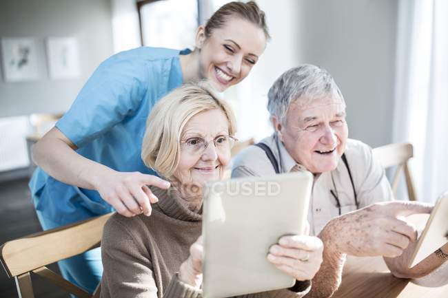 Enfermera sonriente ayudando a pareja mayor a usar tabletas en el hogar de cuidados . - foto de stock