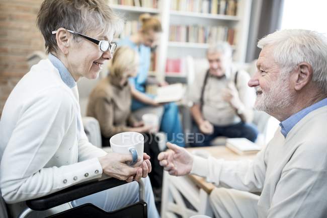 Senior mulher em cadeira de rodas falando com amigo masculino na sala de estar do lar de cuidados . — Fotografia de Stock