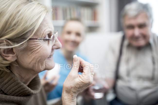 Старшая женщина принимает таблетки в доме, в то время как медсестра и старший мужчина наблюдает
. — стоковое фото