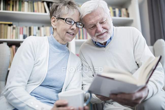Seniorenpaar mit Tasse Tee liest gemeinsam Buch im Sessel. — Stockfoto