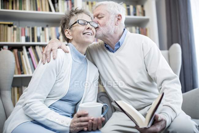Старший мужчина целует женщину в щеку и обнимает во время чтения книги и проведения чая . — стоковое фото