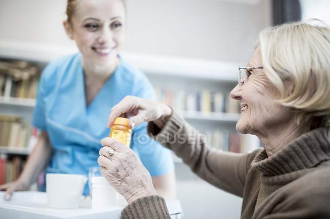 Enfermeira fêmea sorrindo e segurando bandeja enquanto a mulher sênior abrindo frasco de remédio . — Fotografia de Stock