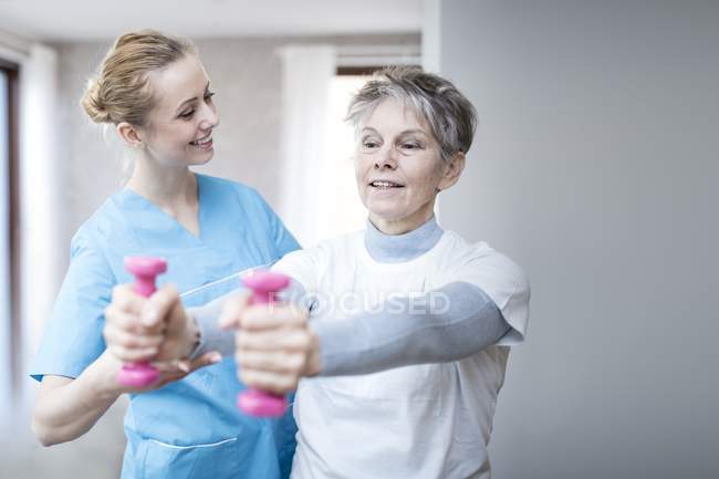 Mulher sênior segurando pesos de mão enquanto fisioterapeuta ajudando . — Fotografia de Stock