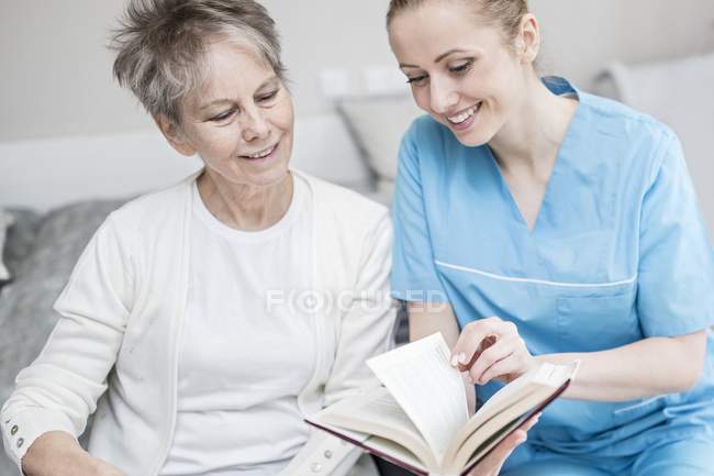 Fröhliche Krankenschwester liest Buch mit Seniorin im Pflegeheim. — Stockfoto
