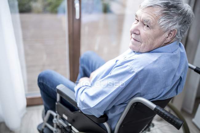 Uomo anziano seduto sulla sedia a rotelle vicino alla finestra in casa di cura . — Foto stock