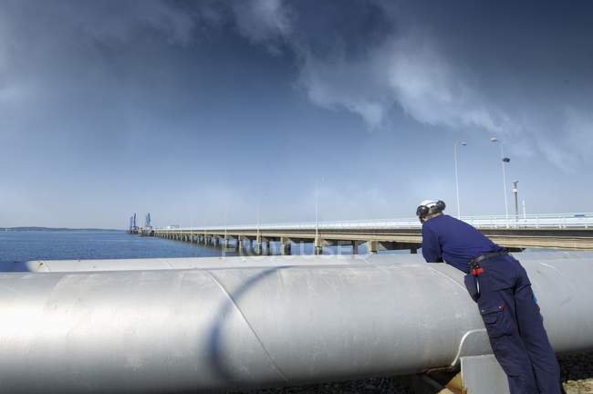 Oil and gas worker leaning on giant pipelines and looking at seascape. — Stock Photo