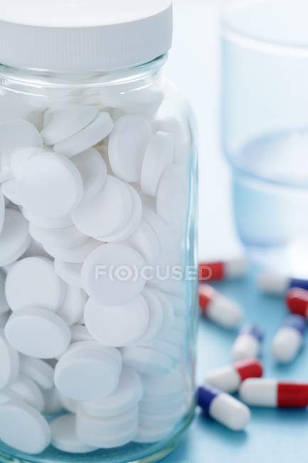Pillole bianche in bottiglia e bicchiere d'acqua con varie capsule in background . — Foto stock