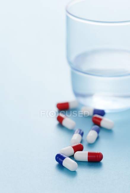 Red and blue drug capsules scattered on blue background with glass of water beside. — Stock Photo