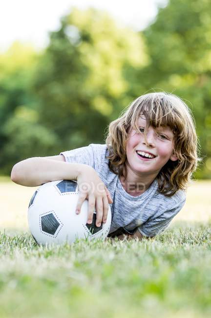 Menino segurando bola de futebol enquanto deitado na grama verde no parque e sorrindo, retrato . — Fotografia de Stock