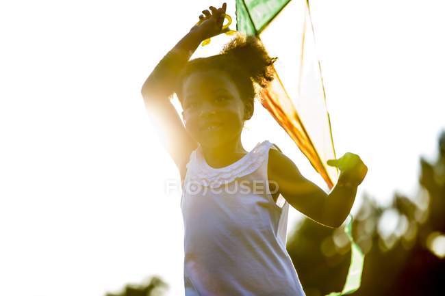 Niña preescolar sosteniendo cometa en el parque y sonriendo con retroiluminación . - foto de stock