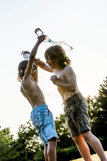 Chicos vertiendo agua el uno sobre el otro de botella de plástico y riendo en el parque . - foto de stock