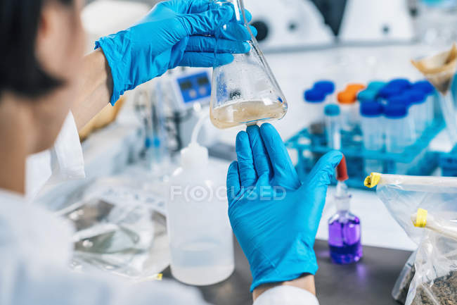 Hands in gloves of female scientist in laboratory shaking glass flask with dissolved samples of soil. — Stock Photo