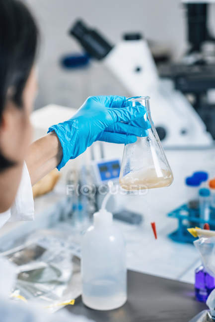 Hände in Handschuhen einer Wissenschaftlerin im Labor schütteln Glaskolben mit gelösten Bodenproben. — Stockfoto