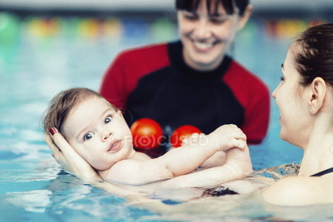 Madre e instructor de natación sumergiendo lentamente al bebé en el agua de la piscina
. - foto de stock