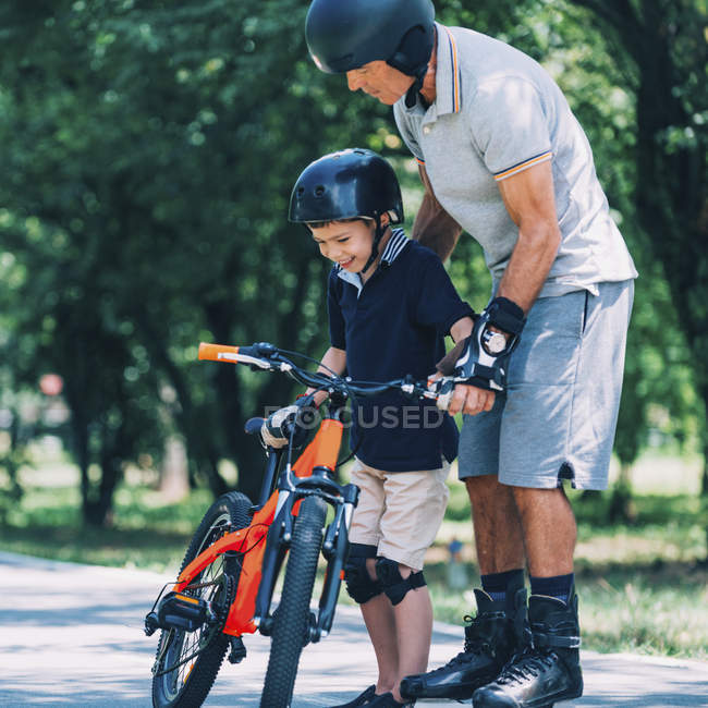 Senior activo en patines enseñando nieto montar en bicicleta en el parque . - foto de stock
