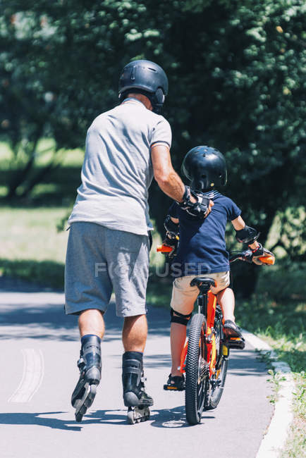 Grandfather and grandson riding together on roller skates and bicycle in park. — Stock Photo