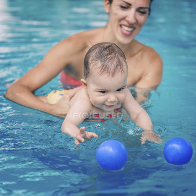 Cute baby boy swimming with mother in pool and trying catching ball. — Stock Photo