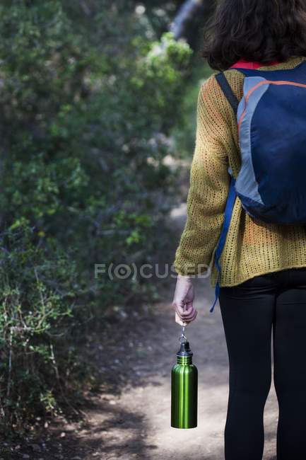 Vue arrière d'une femme marchant sur un sentier forestier tenant une bouteille d'eau . — Photo de stock