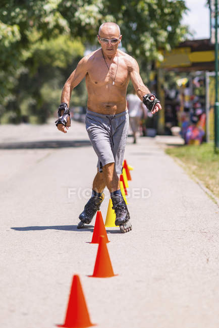 Shirtless senior hombre patinaje sobre ruedas en el parque en la carretera con conos . - foto de stock