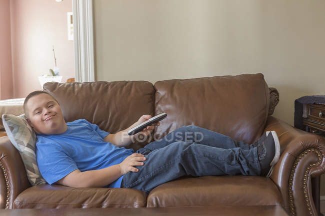 Teenage Boy With Down Syndrome Lying On Couch With Tv Remote