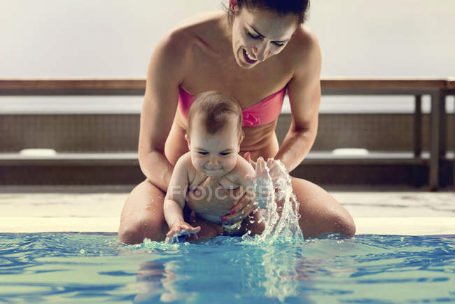 Madre con bambino seduto sul bordo della piscina e spruzzi d'acqua . — Foto stock