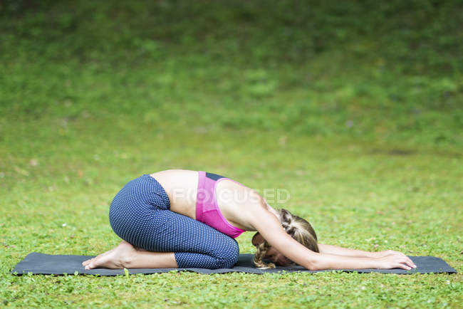Woman practicing half tortoise ardha kurmasana yoga position on mat in park. — Stock Photo