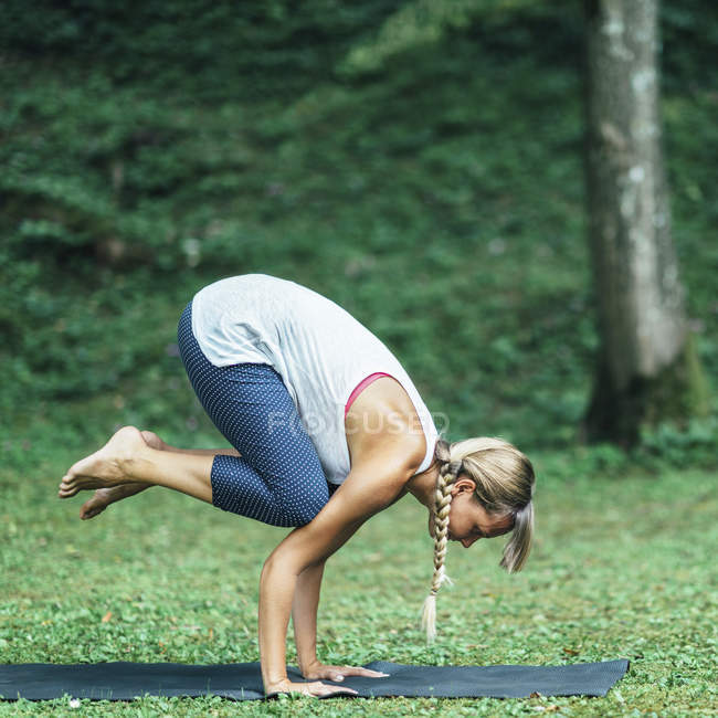 Mujer joven haciendo yoga, practicando la posición de cuervo bakasana en la estera en el parque . - foto de stock