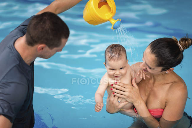 Instructor with baby boy and mother playing in pool and spraying baby with watering can. — Stock Photo
