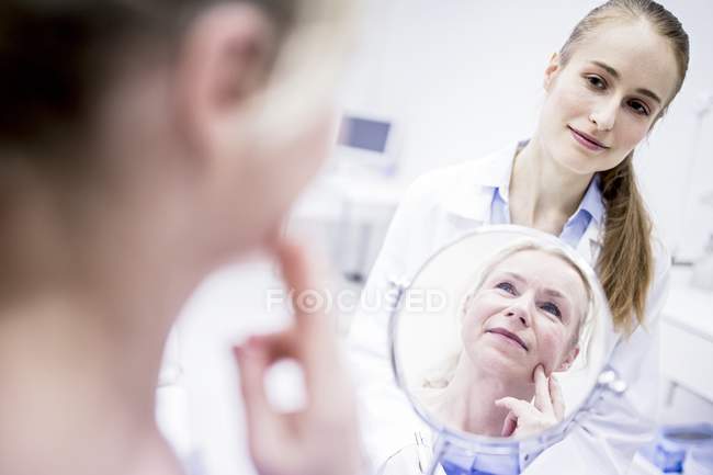 Mulher madura olhando no espelho enquanto médico feminino em pé atrás . — Fotografia de Stock