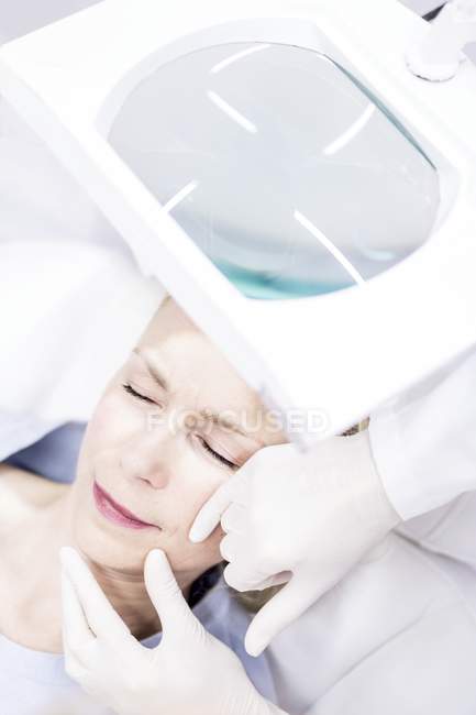 Técnico de beleza examinando a pele facial feminina usando lupa, close-up . — Fotografia de Stock